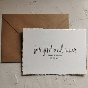 May include: A white card with gold edges and the text "f&uuml;r jetzt und immer" in black cursive writing. Below the text is "Aileen & Ricardo" and the date "07.07.2023".