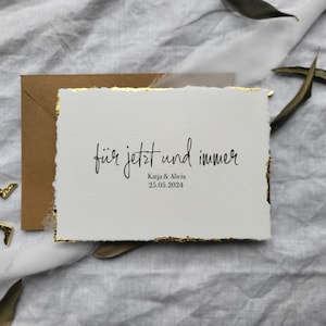 May include: A white card with gold edges and the text "f&uuml;r jetzt und immer" in black script. Below the text is "Katja & Alicia" and the date "25.05.2024".