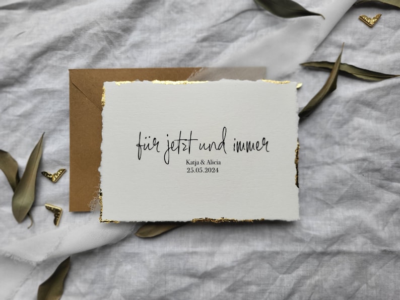 May include: A white card with gold edges and the text "f&uuml;r jetzt und immer" in black script. Below the text is "Katja & Alicia" and the date "25.05.2024".