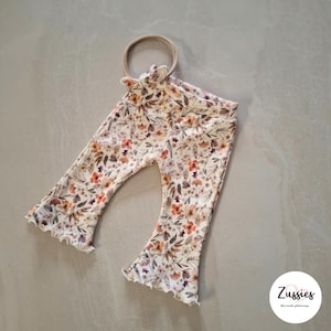 Puede incluir: Pantalones acanalados con dobladillo acampanado y diadema a juego, con un estampado floral en tonos naranja, marr&oacute;n y crema. Los pantalones tienen una cintura el&aacute;stica y un peque&ntilde;o detalle de lazo en la parte delantera. Se ve la marca Zussies.