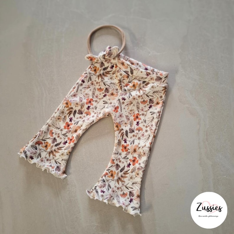 Puede incluir: Pantalones acanalados con dobladillo acampanado y diadema a juego, con un estampado floral en tonos naranja, marr&oacute;n y crema. Los pantalones tienen una cintura el&aacute;stica y un peque&ntilde;o detalle de lazo en la parte delantera. Se ve la marca Zussies.