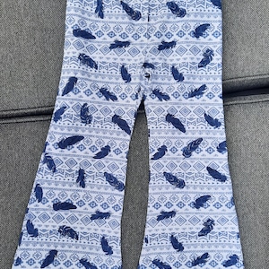 Puede incluir: Un pantal&oacute;n de campana blanco con un patr&oacute;n repetitivo de plumas azul oscuro y formas geom&eacute;tricas. El pantal&oacute;n tiene un dise&ntilde;o de cintura alta con una silueta de pierna ancha y acampanada que se ensancha notablemente desde la rodilla al bajo.