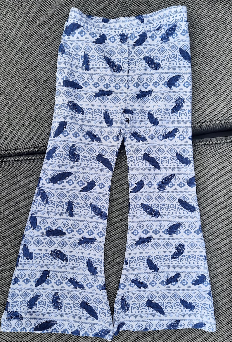 Puede incluir: Un pantal&oacute;n de campana blanco con un patr&oacute;n repetitivo de plumas azul oscuro y formas geom&eacute;tricas. El pantal&oacute;n tiene un dise&ntilde;o de cintura alta con una silueta de pierna ancha y acampanada que se ensancha notablemente desde la rodilla al bajo.