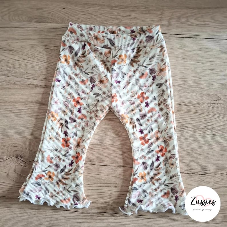 Puede incluir: Pantalones acampanados de canal&eacute; en color blanco roto con un estampado floral de hojas y flores naranjas, marrones y moradas. Los pantalones tienen una cintura el&aacute;stica y dobladillos con volantes. Se ve un logotipo circular blanco con el texto Zussies.