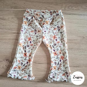 Op de afbeelding: Witte flared leggings met een bloemenprint met oranje, bruine en paarse bloemen. De leggings hebben een gerimpelde zoom.