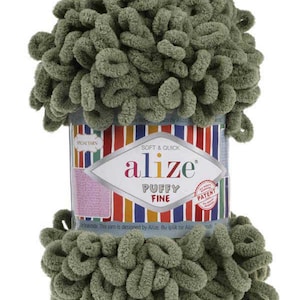 Puede incluir: Ovillo de lana Alize Puffy Fine en color verde apagado. La lana está en bucles, creando una apariencia suave y texturizada. La etiqueta presenta el nombre de la marca y los detalles del producto, con un diseño a rayas colorido.