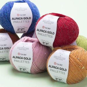 May include: Close-up of several balls of Alpaca Gold Paillettes yarn in various colors, including blue, red, pink, and orange. Each ball has a white label with the brand name and product details. The yarn appears soft and textured.