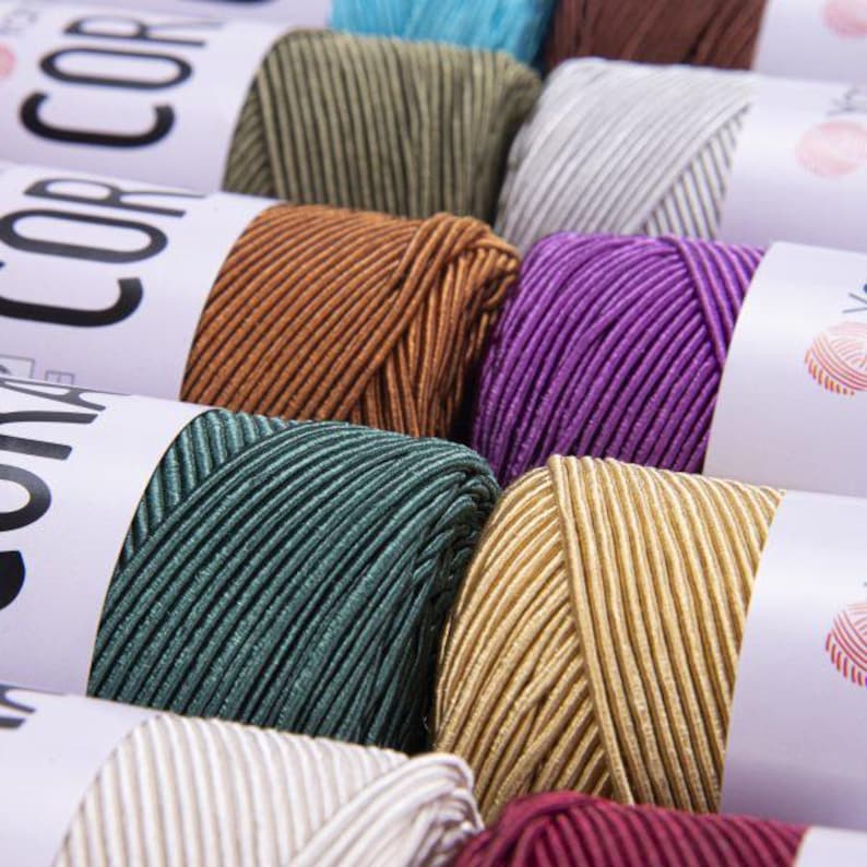 Puede incluir: Primer plano de carretes de hilo en varios colores, incluyendo verde azulado, dorado, morado, marr&oacute;n y blanco. El hilo es texturizado y tiene un brillo sutil.