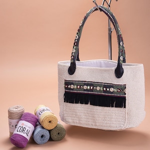 Puede incluir: Un bolso tote blanco de crochet con flecos negros y un ribete de cuentas. El bolso tiene un asa de cuero negro y est&aacute; sobre una superficie marr&oacute;n claro. Hay cinco ovillos de hilo de diferentes colores junto al bolso.
