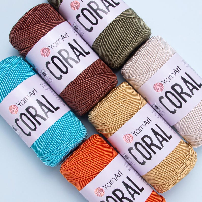Puede incluir: Seis madejas de hilo en diferentes colores, incluyendo turquesa, naranja, beige, marr&oacute;n, verde y marr&oacute;n oscuro. Cada madeja tiene una etiqueta blanca con el texto "YarnArt CORAL" impreso en ella.