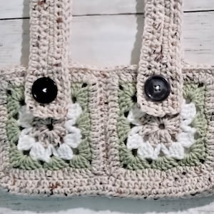 Op de afbeelding: Een gehaakte draagtas met twee grote vierkanten in het midden. De vierkanten zijn gemaakt van wit en groen garen en hebben een bloemenpatroon. De tas heeft twee beige hengsels en zwarte knopen.