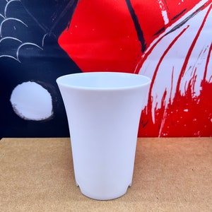 May include: A white ceramic cup with a rounded bottom and a slightly flared lip. The cup is sitting on a brown surface.