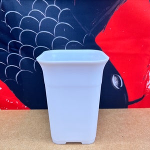 May include: A small, white, square-shaped planter with a slightly flared top. The planter is made of a smooth, matte material and sits on a flat base. The background features a dark blue and red abstract design.