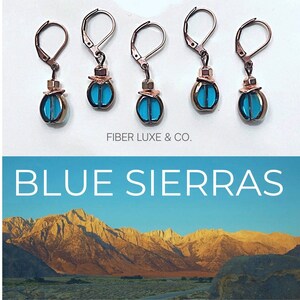 May include: Five pairs of earrings with copper hooks and blue glass teardrop-shaped charms. The earrings are arranged in a row on a white surface. The text "FIBER LUXE & CO." is below the earrings. The text "BLUE SIERRAS" is below the earrings and the text. The background is a photo of a mountain range.