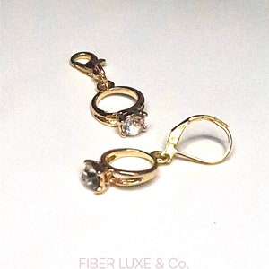 May include: Two gold-colored ring charms with clear, round gemstones. One charm has a lobster clasp, and the other has a lever back earring hook. The charms are on a white background. The text "FIBER LUXE & Co." is at the bottom.