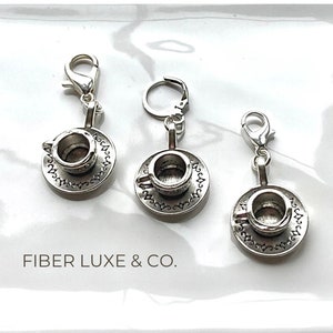 May include: Three silver charm pendants in the shape of a teacup and saucer. Each charm has a silver loop for attaching to a bracelet or necklace. The charms are on a white background.