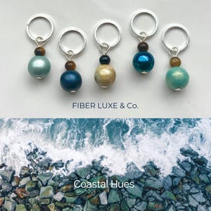 May include: Five stitch markers with silver rings and colorful glass beads. The beads are teal, blue, beige, blue, and teal. The text "FIBER LUXE & Co." is below the stitch markers. The text "Coastal Hues" is below the stitch markers and a photo of ocean waves crashing on rocks.