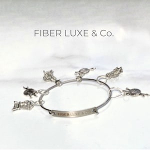 May include: A silver bangle bracelet with charms shaped like balls of yarn and knitting needles. The bracelet has an engraved tag that reads "Fiber Luxe & Co."