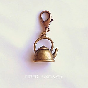 May include: A gold-toned metal teapot charm with a loop for attaching to a bag or keychain. The charm is a miniature replica of a traditional teapot with a lid, spout, and handle.