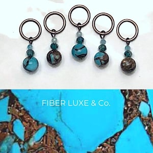 Puede incluir: Conjunto de cinco marcadores de punto turquesa y cobre con una cuenta de piedra turquesa y marrón en cada uno. Los marcadores de punto tienen forma de círculo y tienen un pequeño bucle en la parte superior. Las cuentas son todas de diferentes formas y tamaños. El texto "FIBER LUXE & Co." es visible en la imagen.