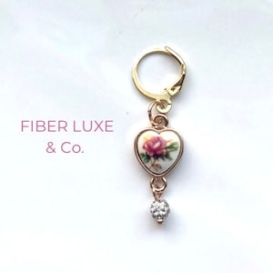 May include: Gold charm with a heart-shaped porcelain pendant featuring a pink rose design. The charm is attached to a gold ring and a small clear crystal dangle.