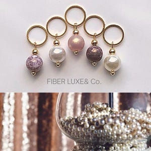 Puede incluir: Seis marcadores de punto de oro con cuentas de diferentes colores. Las cuentas son rosadas, blancas y de color púrpura y blanco moteado. El texto "FIBER LUXE & Co." está impreso debajo de los marcadores de punto.