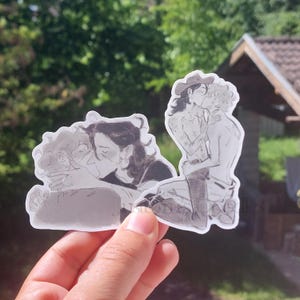 以下が含まれることがあります： 親密な瞬間を描いたグレースケールイラストのステッカーセット。1つは2人の人物が抱き合っている様子、もう1つはカウボーイハットをかぶったカップルがキスをしている様子です。ステッカーは手で持たれています。