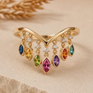 Puede incluir: Un anillo dorado con un diseño en forma de V, adornado con gemas colgantes de colores en tonos azul, amarillo, verde, rosa, naranja y morado. Pequeñas piedras claras están engastadas entre las gemas.