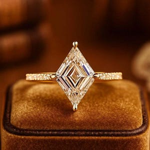 May include: A gold engagement ring featuring a large, kite-cut diamond. The diamond is set in a gold band with small accent stones. The ring is presented on a brown velvet ring box.