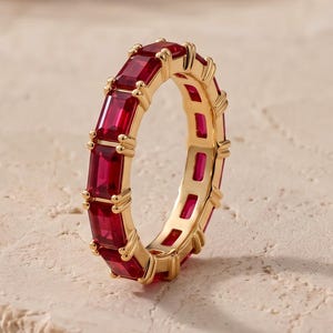 Puede incluir: Un anillo de eternidad en oro con una hilera continua de gemas de color rojo vibrante, talladas en corte esmeralda y sujetas con garras. El anillo presenta un acabado metálico pulido y un diseño uniforme que rodea toda la circunferencia.