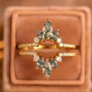 May include: A gold ring set featuring two bands with marquise-cut moss agate stones and small round clear accent stones. The design includes a pointed, crown-like arrangement of green-flecked moss agate gems set in yellow gold metal bands.