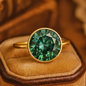 May include: A gold ring with a large, round, emerald-green gemstone. The gemstone is set in a gold bezel, and the ring is displayed on a brown velvet ring box. The ring's band is a simple, polished gold.