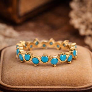 May include: A gold-colored ring with a repeating pattern of small, round, turquoise-colored stones. The ring is set on a brown velvet surface, with a blurred background. The ring is a delicate piece of jewelry.
