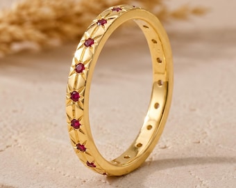 Puede incluir: Un anillo de banda dorada que presenta una serie de pequeños rubíes rojos redondos engastados dentro de patrones de estrellas grabados que rodean la superficie exterior del metal, creando un diseño repetitivo.
