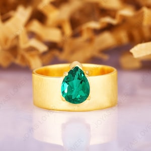 Puede incluir: Un anillo dorado con una banda ancha y una piedra preciosa verde esmeralda en forma de pera. El anillo está sobre un fondo borroso con el texto "JOYAS LOVER" repetido.