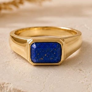 May include: A gold-toned ring featuring a rectangular, deep blue lapis lazuli gemstone set in the center. The band is smooth and polished, with a flat, bezel-style setting that holds the stone securely. The design is clean and symmetrical.