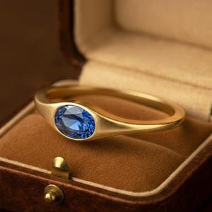 May include: A gold ring with an oval, blue gemstone, displayed in a brown velvet-lined jewelry box. The ring has a smooth, polished band and a bezel setting for the gemstone. The box is closed with a small, gold-colored clasp.