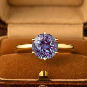 Può includere: Un anello in oro con una pietra preziosa rotonda e sfaccettata dai toni viola e blu, sostenuta da una montatura a sei griffe. L'anello è esposto all'interno di un portagioie rettangolare marrone foderato in velluto con una chiusura dorata.