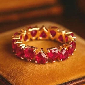 Könnte beinhalten: Ein goldener Eternity-Ring mit einer durchgehenden Reihe leuchtend roter, birnenförmiger Edelsteine. Der Ring ist mit einer polierten Metallfassung versehen, die jeden tropfenförmigen Stein sicher hält und ein kreisförmiges Design in Rot bildet.