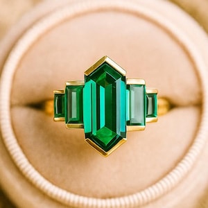 Puede incluir: Un anillo de oro con una gran piedra verde talla esmeralda en el centro, flanqueada por piedras verdes rectangulares más pequeñas. El anillo está engastado en una banda de oro y descansa en un joyero beige.