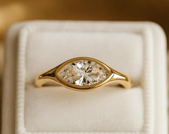 May include: A gold ring featuring a marquise-cut clear gemstone set in a smooth bezel setting. The ring is displayed centred in a white velvet ring box, highlighting the polished gold band and the faceted stone's elongated shape.