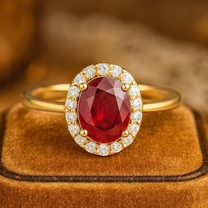 Può includere: Anello in oro con una pietra centrale ovale di rubino, circondata da piccoli diamanti. L'anello è montato su una fascia d'oro e viene presentato su una superficie di velluto marrone.