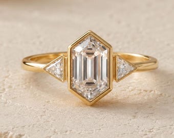May include: A gold ring featuring a central hexagonal-cut diamond flanked by two smaller triangular-cut diamonds. The band is crafted from polished yellow gold, showcasing a clean, geometric design with clear, faceted stones set in a metal frame.