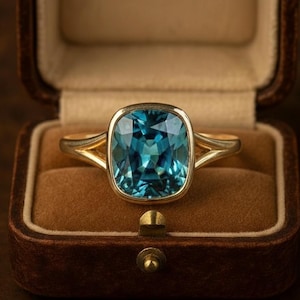 May include: A gold ring featuring a large, teal-coloured, square-cut gemstone. The ring is set in a gold band and presented in an open, brown velvet ring box. The box highlights the ring's features.