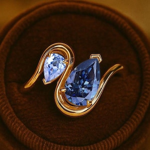 Può includere: Un anello d'oro con due pietre preziose blu a forma di goccia. L'anello è incastonato in un portagioie di velluto marrone, con una fascia d'oro che si curva attorno a ogni pietra. L'anello è esposto su un piatto marrone con un centrino di pizzo bianco.