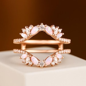 May include: Rose gold ring guard with a double band design. The ring features a halo of small, clear stones and larger, pear-shaped opals. The opals are a soft pink color, and the ring is set on a white surface.