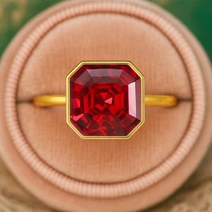 Op de afbeelding: Een gouden ring met een grote, vierkant geslepen robijn. De robijn is gezet in een gouden bezel, en de ring wordt gepresenteerd in een roze fluwelen ringdoos. De achtergrond is zachtgroen en goud.