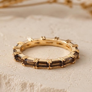 May include: A gold band ring featuring a continuous row of rectangular, baguette-cut smoky brown gemstones set in a prong setting. The ring has a polished metallic finish and a uniform design that encircles the entire circumference of the band.