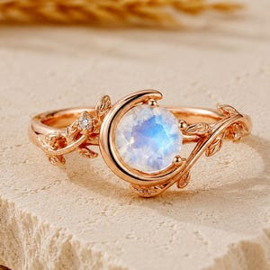 Puede incluir: Un anillo de oro rosa con una piedra lunar redonda e iridiscente en el centro, engastada en un marco de media luna. La banda presenta motivos detallados de hojas y enredaderas, con una pequeña piedra preciosa clara en un lado.