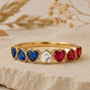 May include: A gold ring featuring a row of seven heart-shaped gemstones. The stones are arranged in a sequence of two blue sapphires, one clear white diamond, and two red rubies, all set within a polished gold band with a smooth finish.